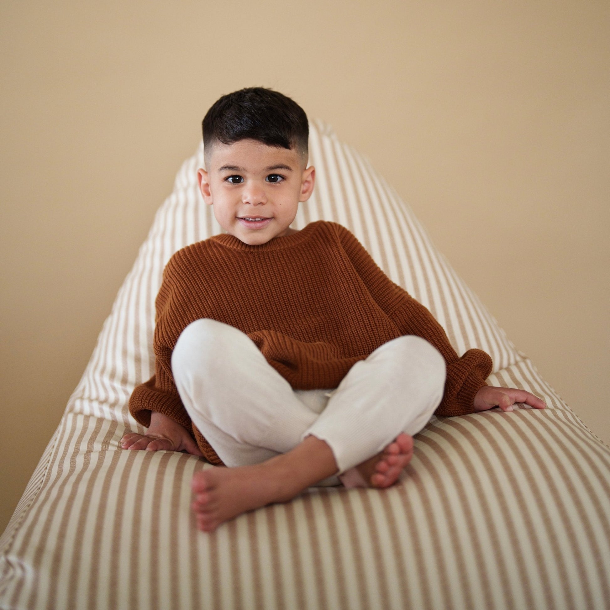 Child sitting on a striped bean bag chair wearing a brown sweater and white pants.