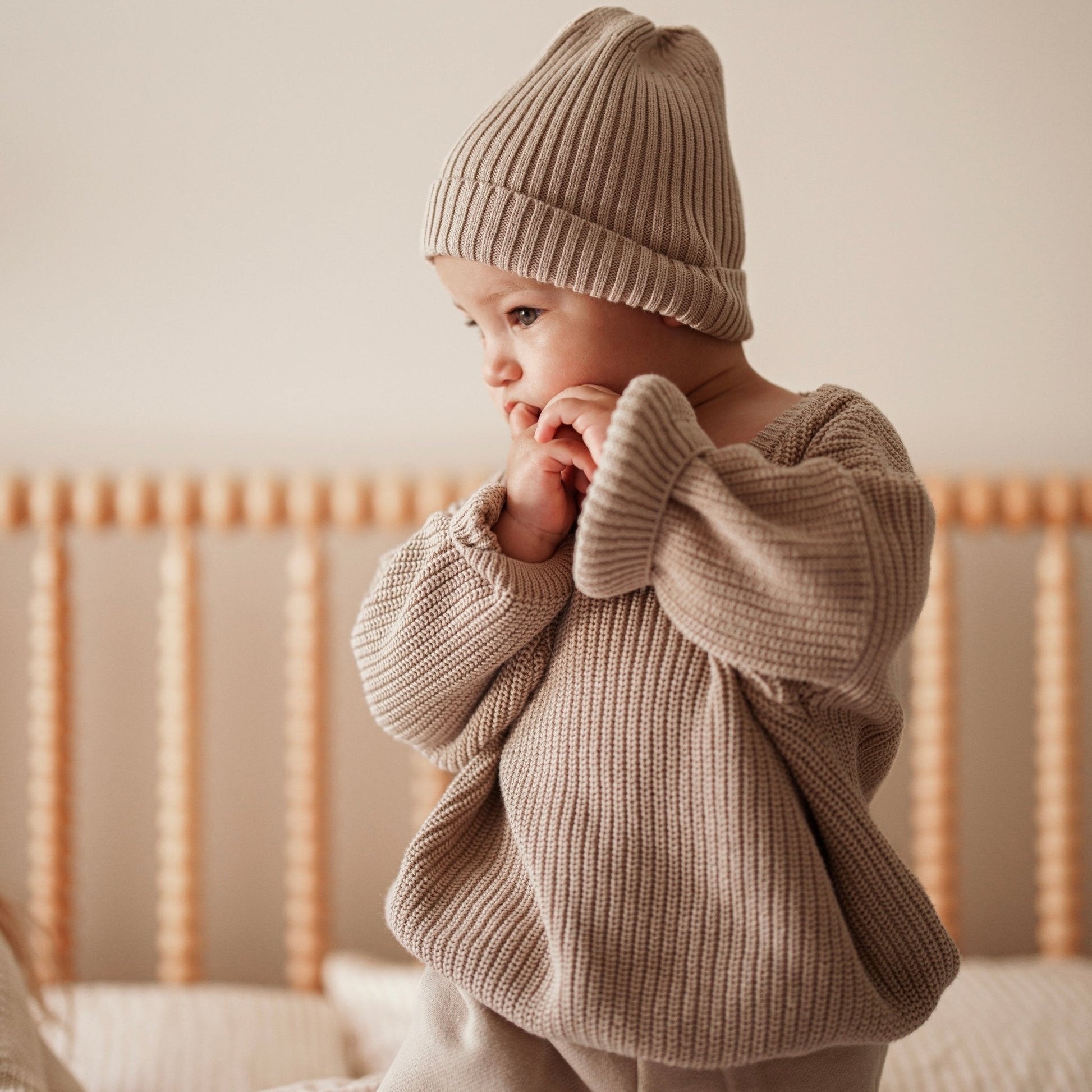 Baby wearing a beige knit outfit and hat, standing in a crib.