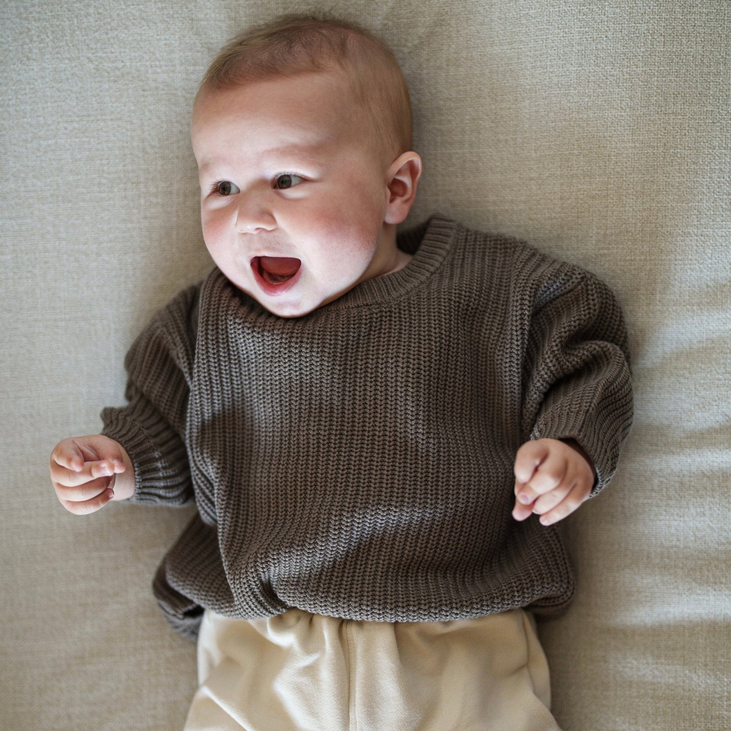 Baby wearing a brown sweater and beige pants lying on a beige surface