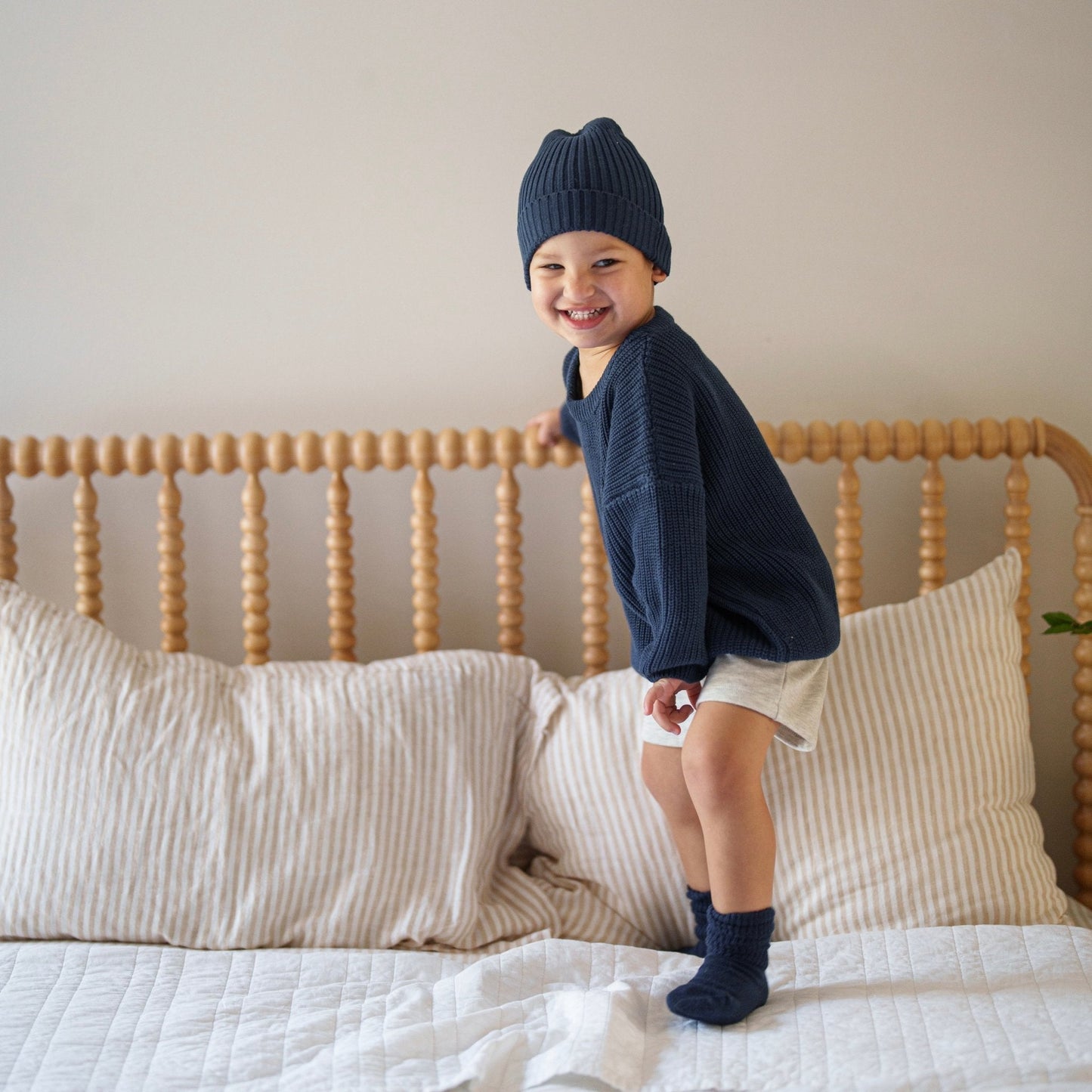 Child wearing a navy sweater and hat standing on a bed with wooden headboard and beige pillows.