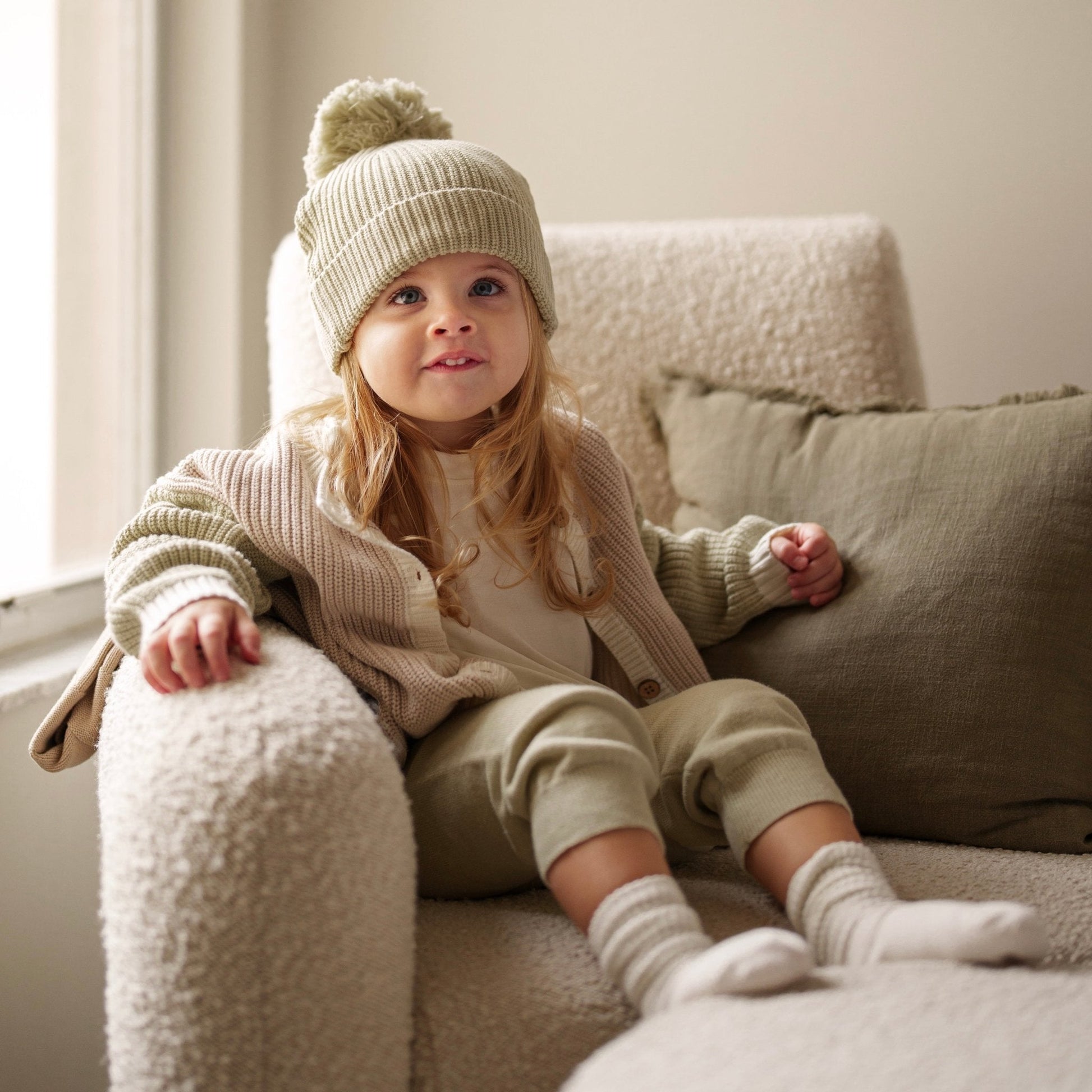 Child wearing a cozy outfit with a knit hat, sweater, and pants sitting on a soft chair.