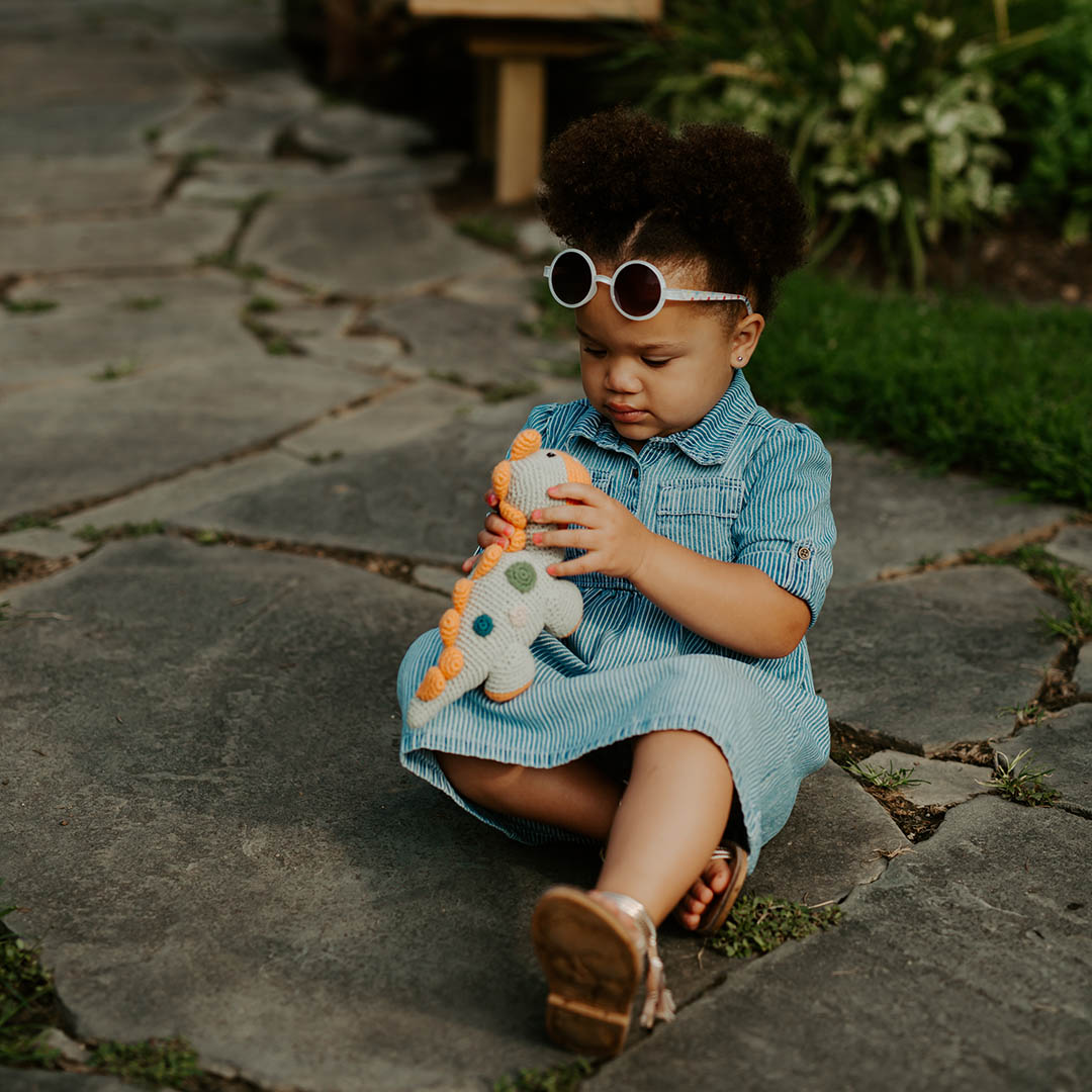 Child in a blue dress sitting on stone steps holding a stuffed dinosaur