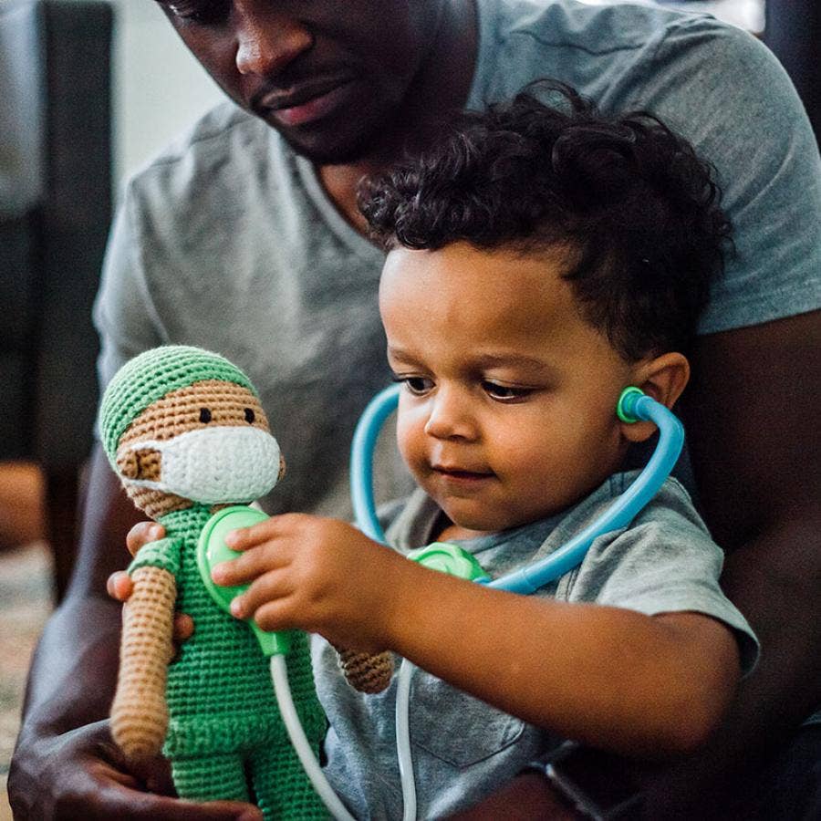 Child playing with a green toy doctor doll and stethoscope, held by an adult.