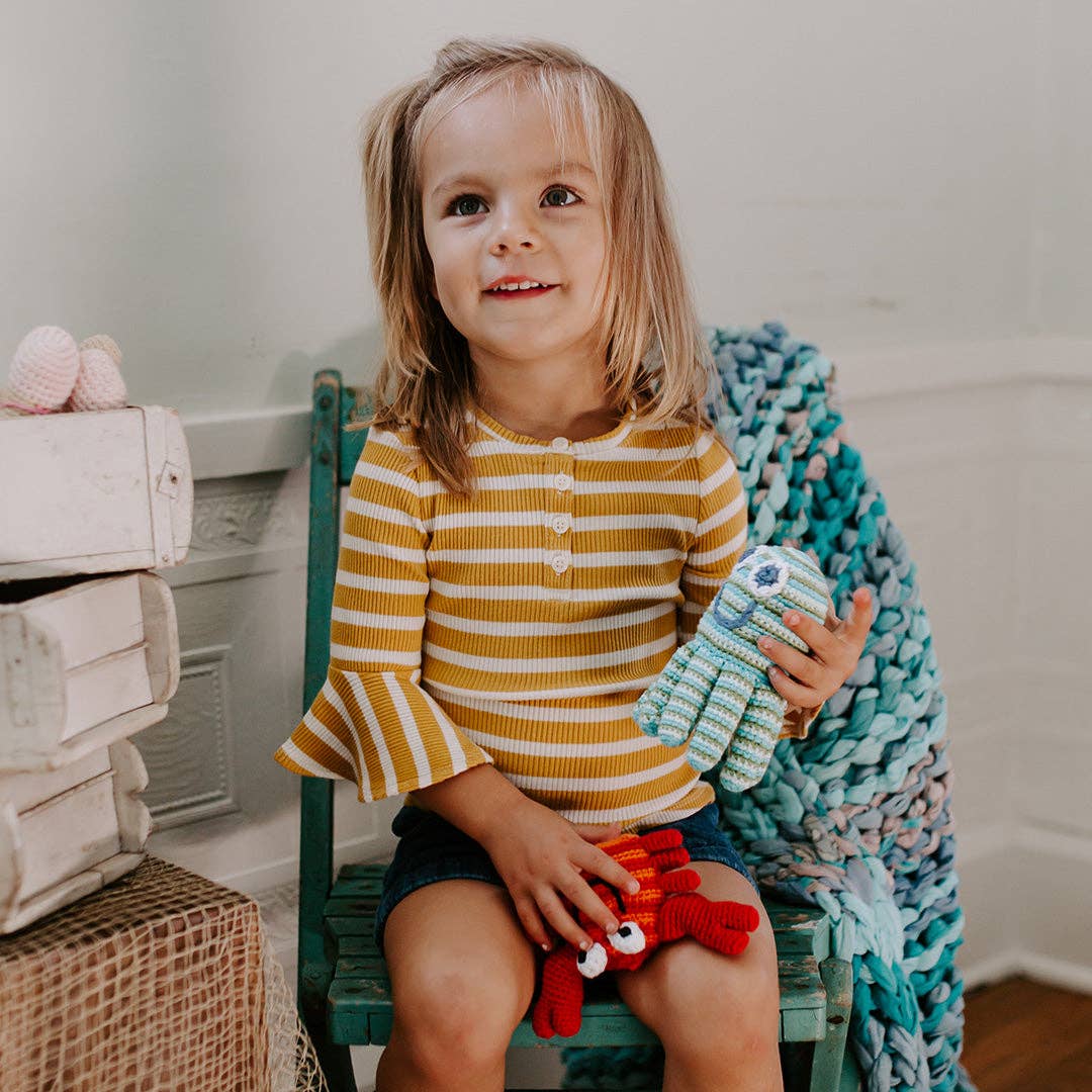 Child sitting on a chair holding a knitted octopus toy, with a cozy blanket and toys in the background.