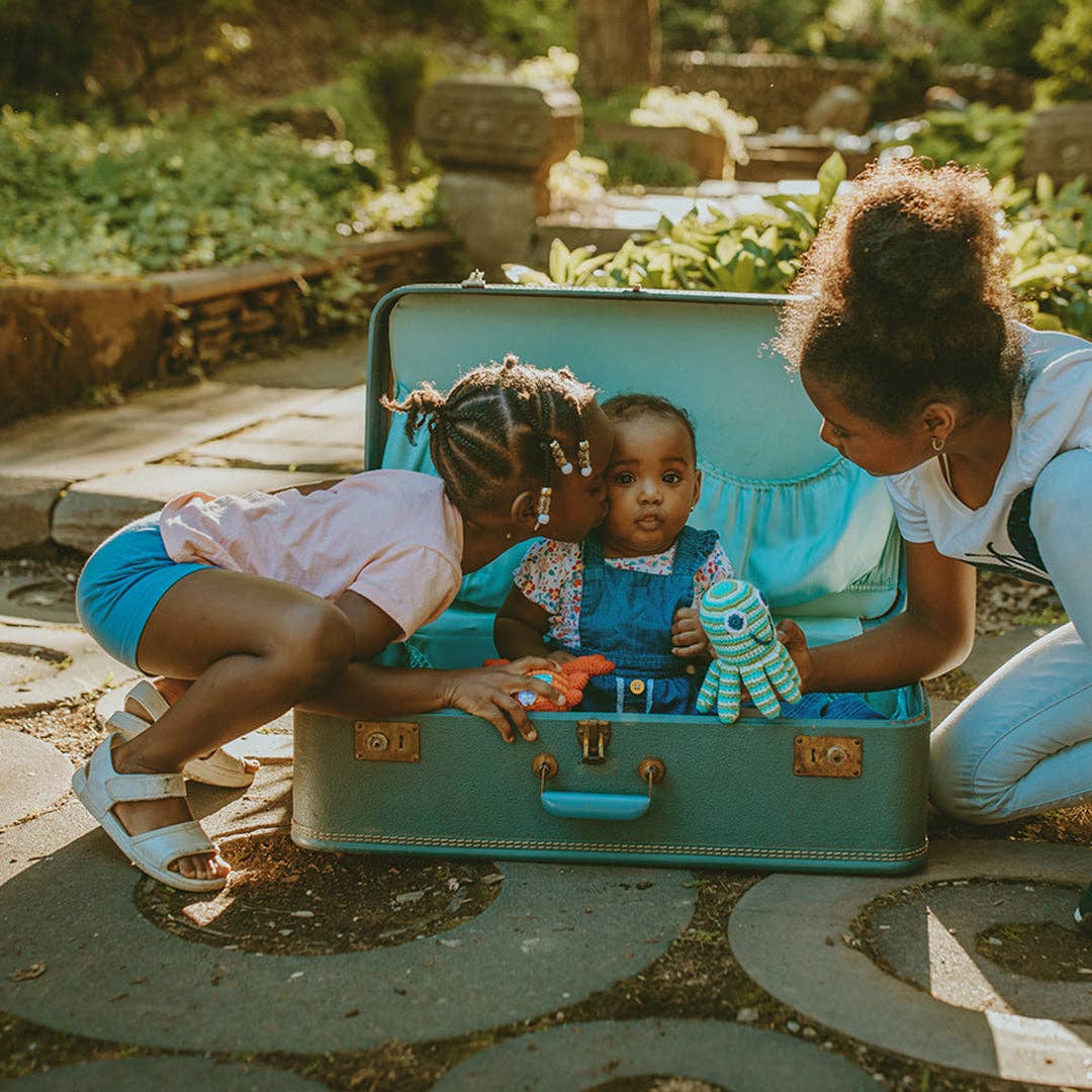 Two children playing inside a teal suitcase with a woman outdoors.