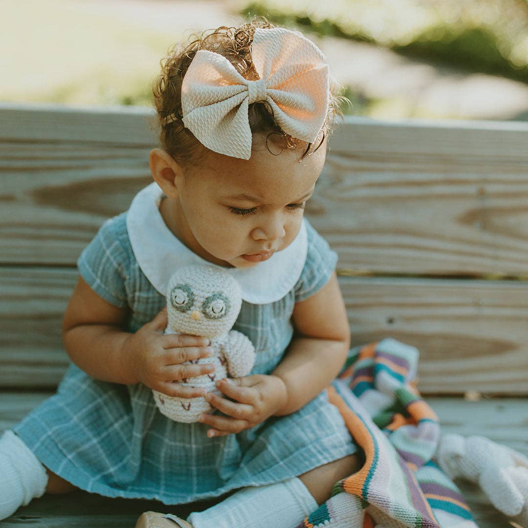 baby wearing a large bow headband and a dress. holding a stuffed owl toy outdoors.