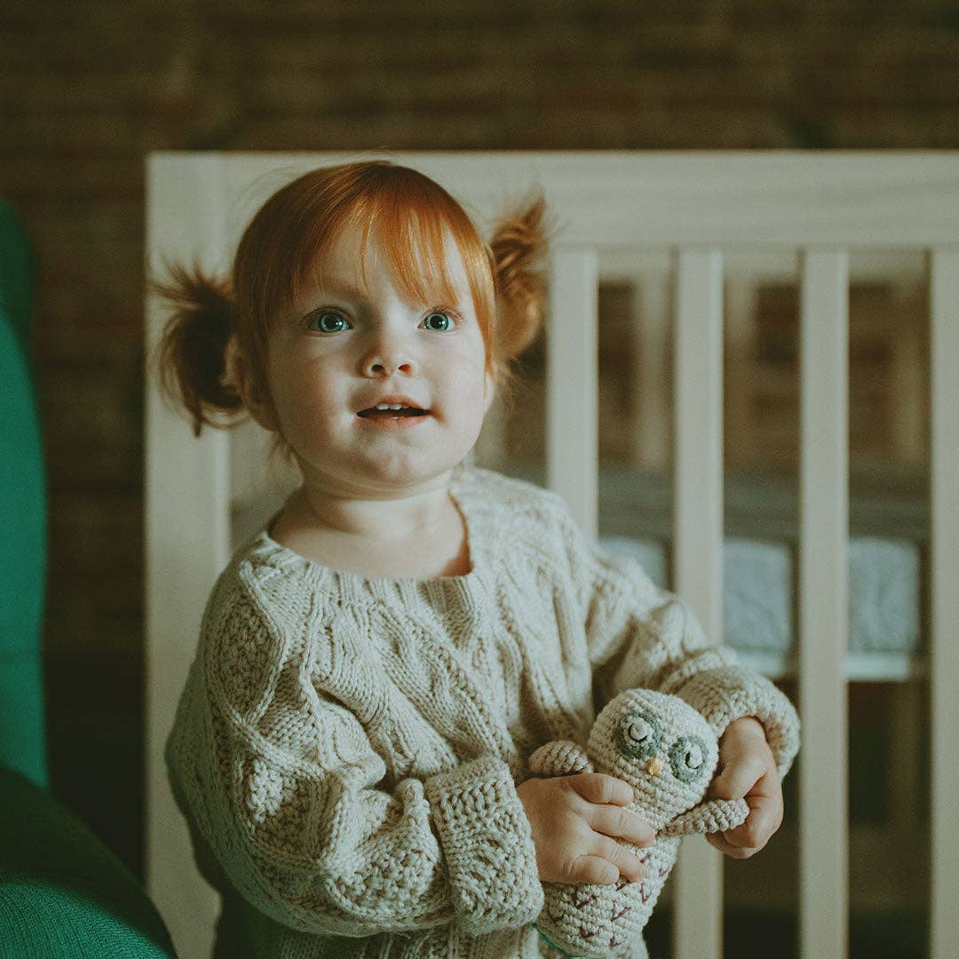 Child in a knitted sweater holding an owl toy in front of a crib