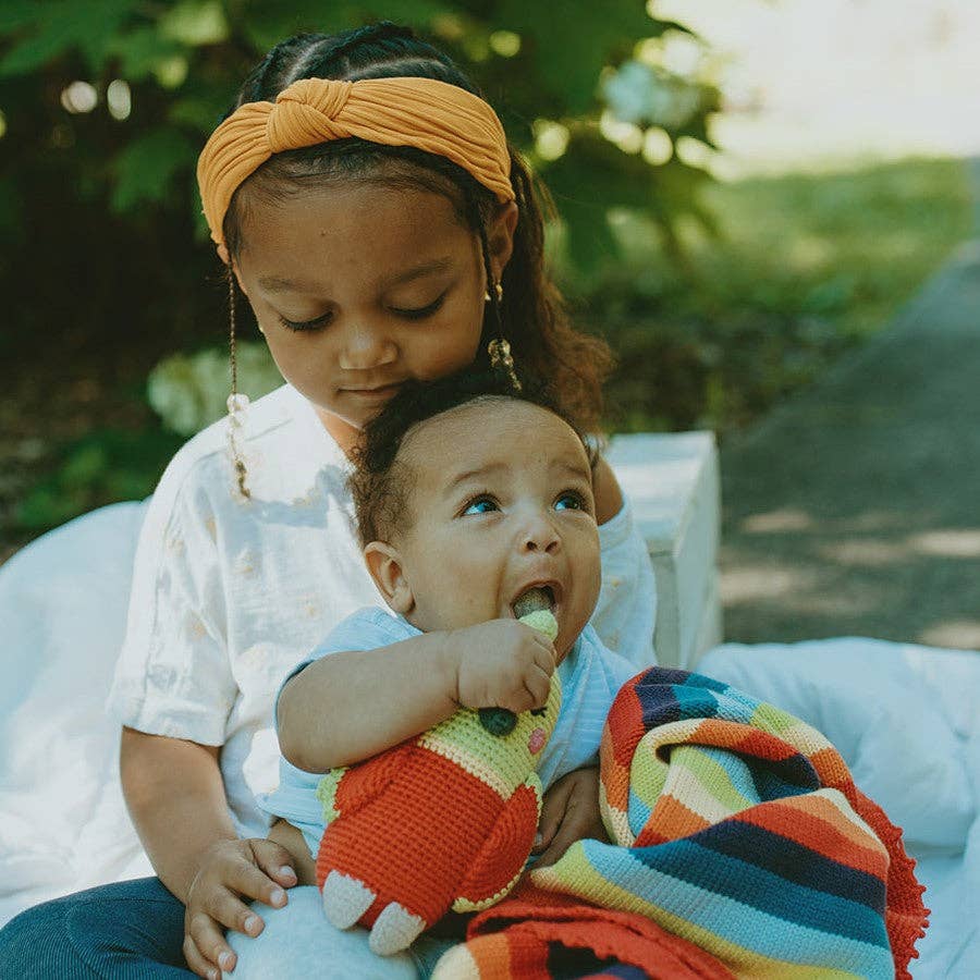 Two children sitting outdoors with a rainbow blanket and toy.