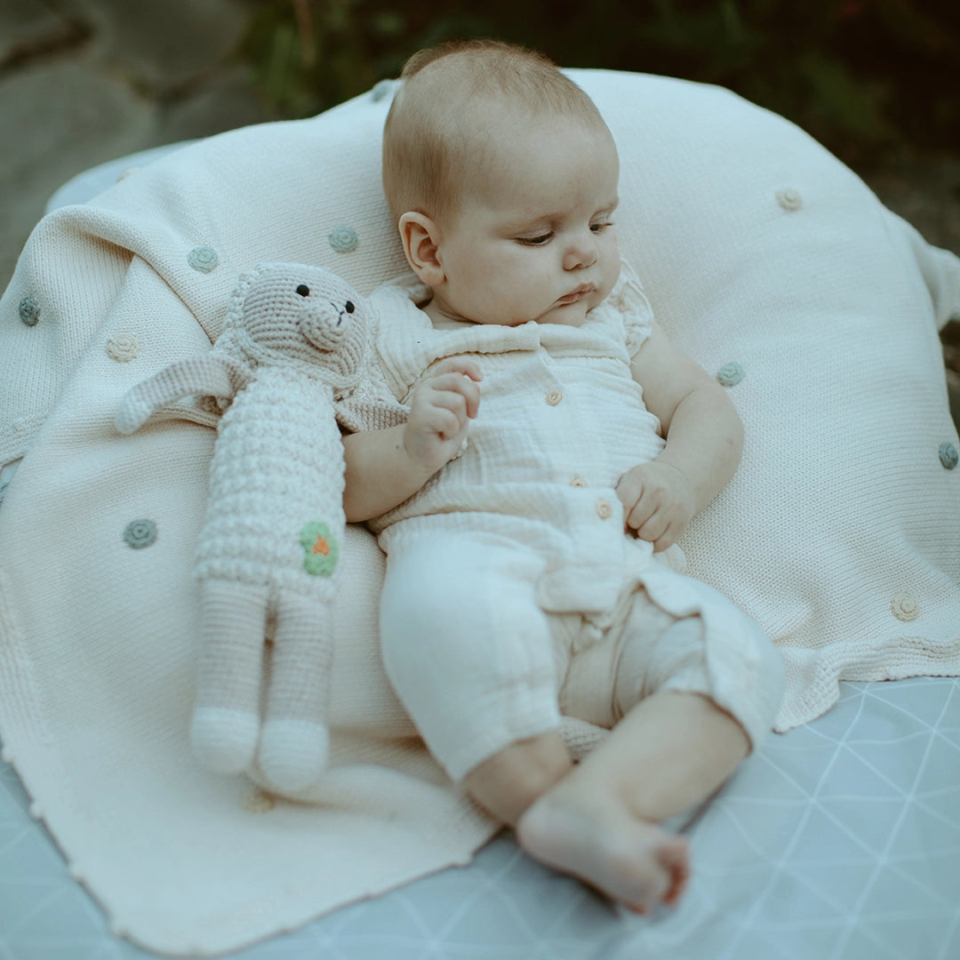 Baby in a white outfit holding a knitted toy on a soft spotty blanket