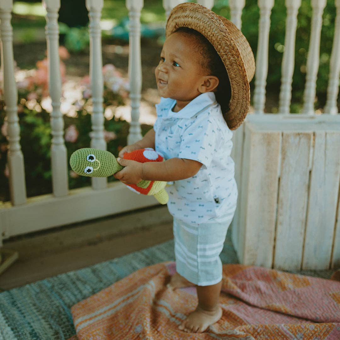 Child wearing a straw hat and light blue outfit, standing on a colorful rug with a garden background holding a turtled knitted stuffed toy