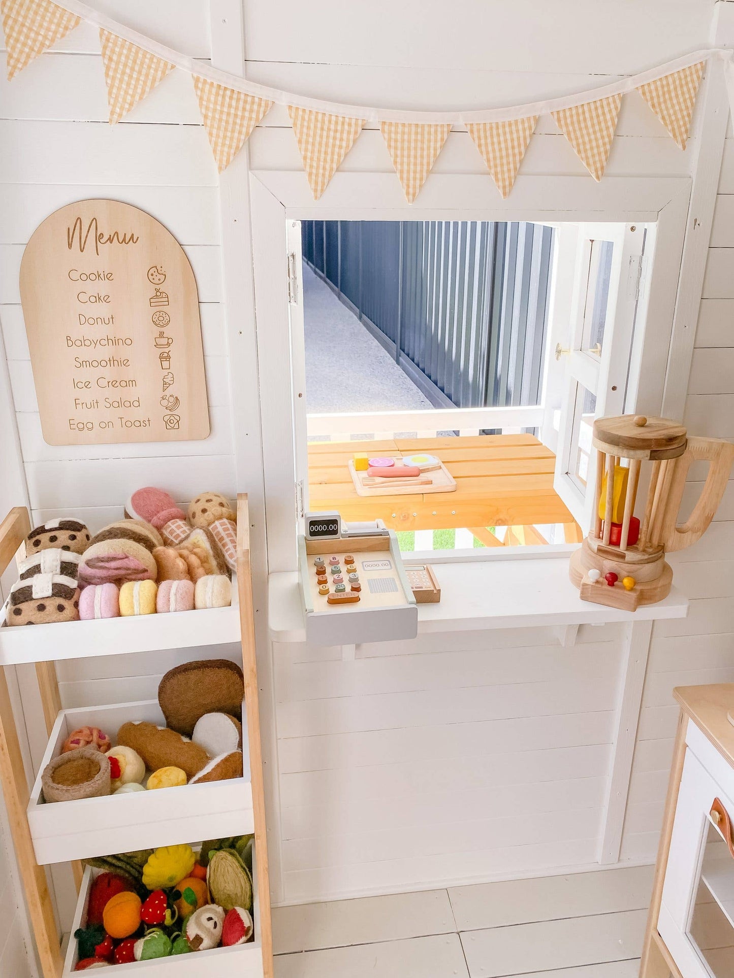 a play coffee shop with felt baked goods and fruits in a shelf and a wooden cash register and blender on thee windowsill