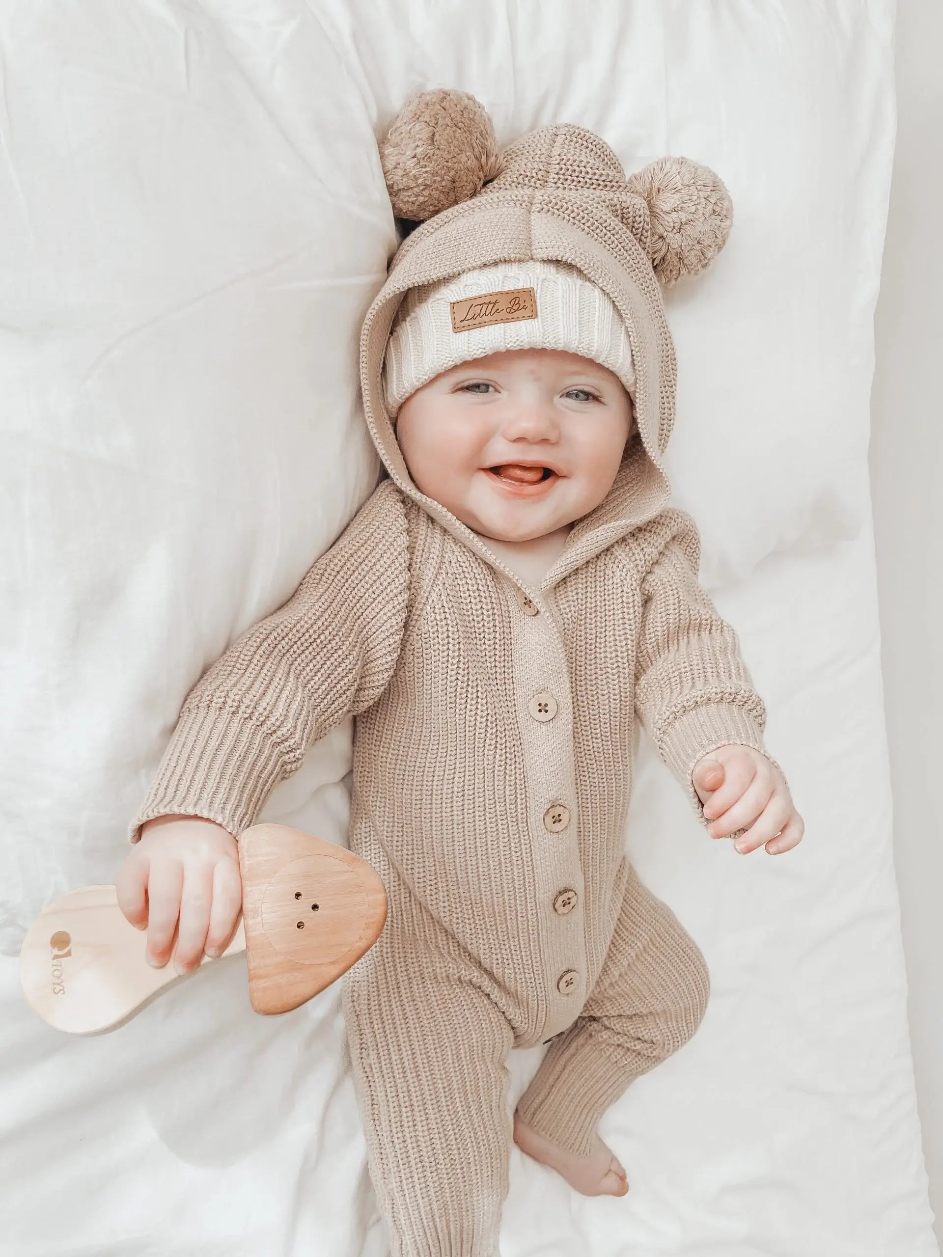 a baby laying on a bed wearing a hooded romper with bear ears holding a wooden mushroom rattle