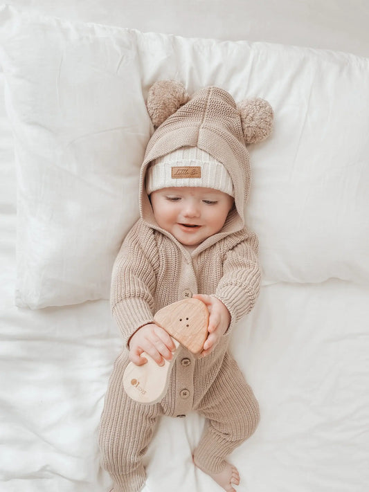 a baby laying on a bed wearing a hooded romper with bear ears holding a wooden mushroom rattle