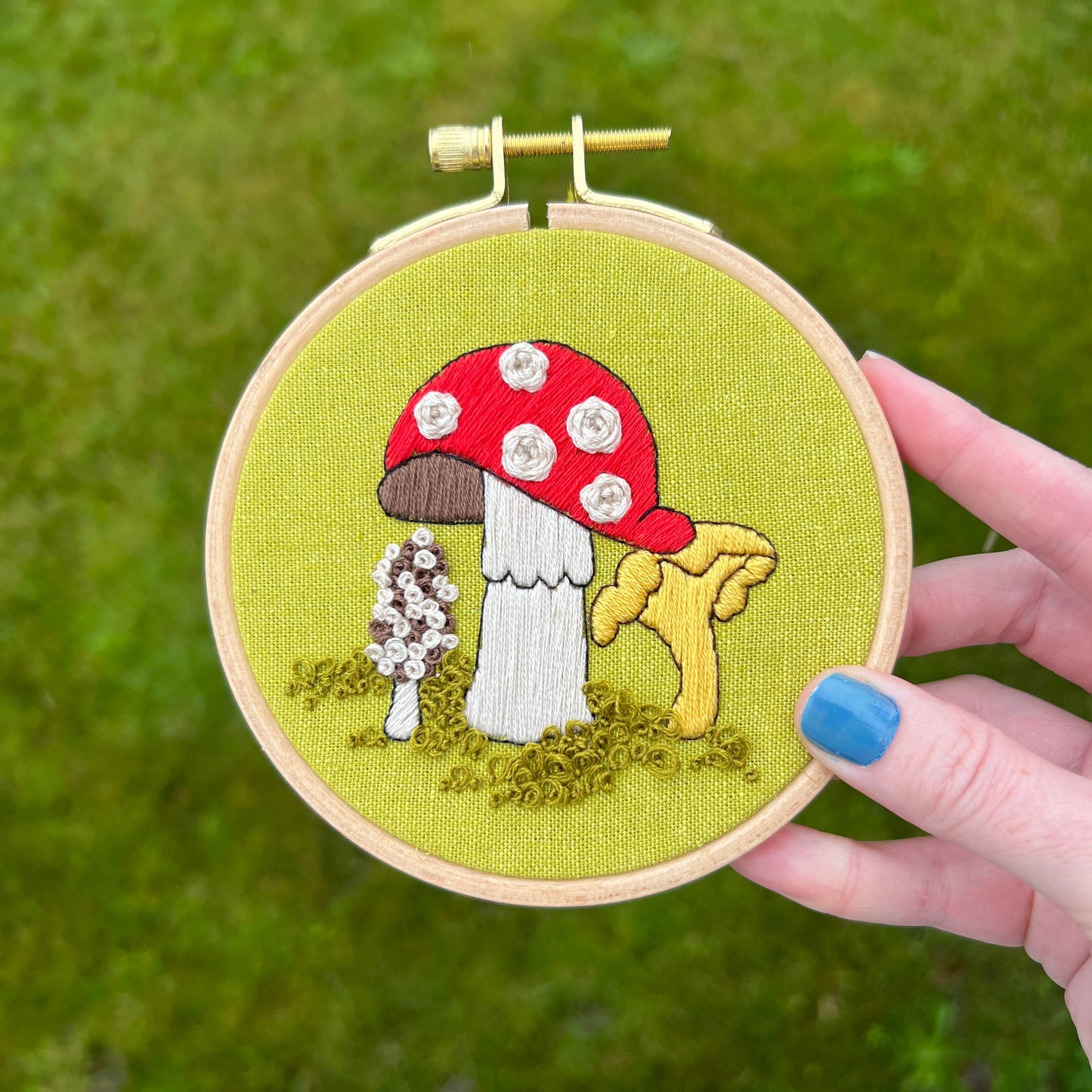 Embroidery hoop with embroidered mushrooms held by a hand against a grass background