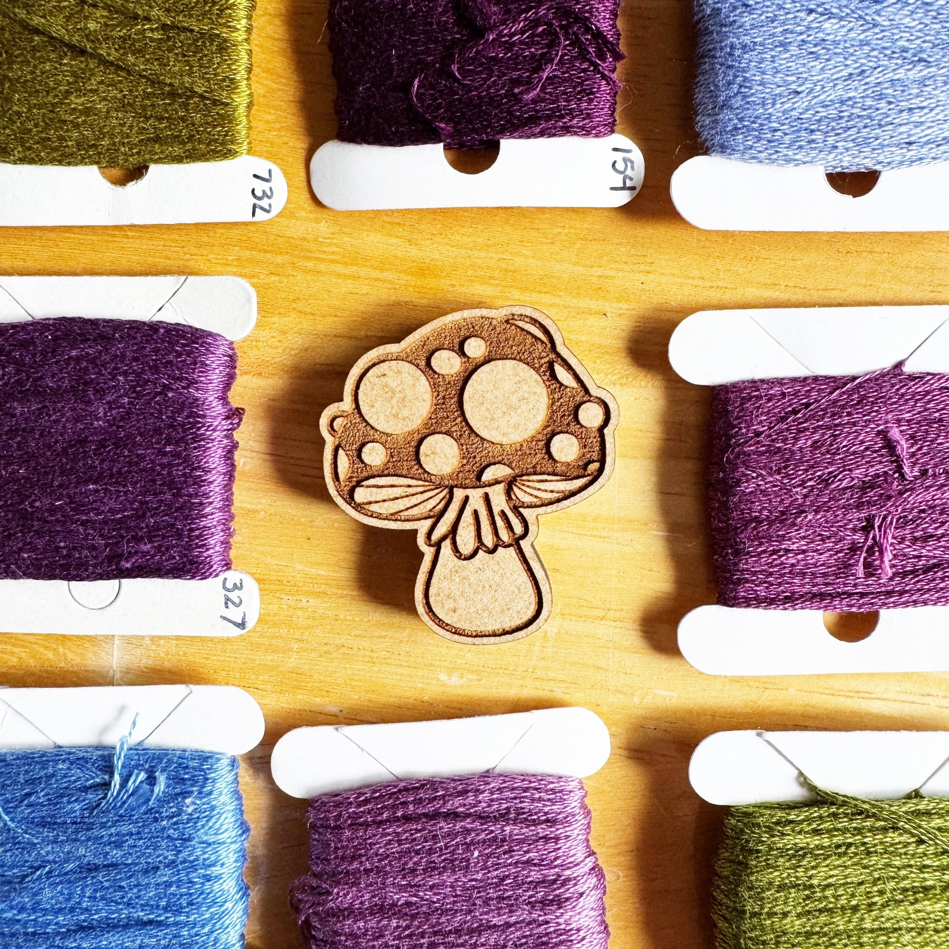 Wooden mushroom-shaped needleminder surrounded by spools of colorful embroidery thread on a wooden board.
