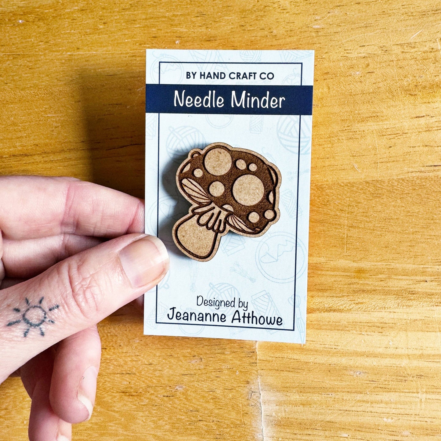 Wooden mushroom-shaped needle minder on a By Hand Craft Co card held by a hand on a wooden surface