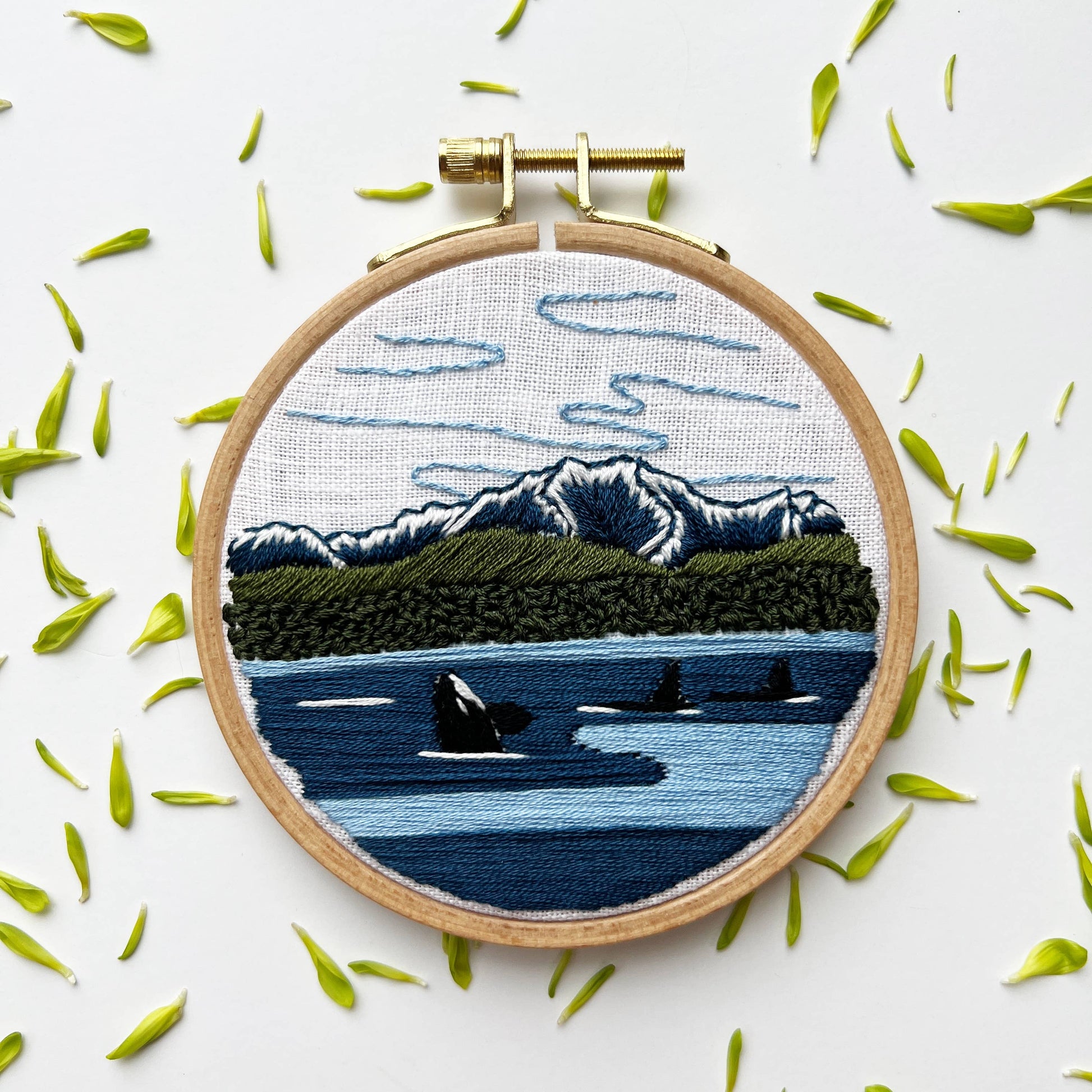 Embroidery hoop with a scenic landscape design featuring mountains, water, and orcas on a white background.