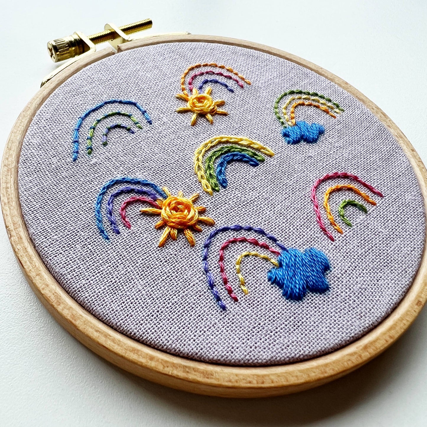 Embroidery hoop with colorful rainbows, suns, and clouds on gray fabric.