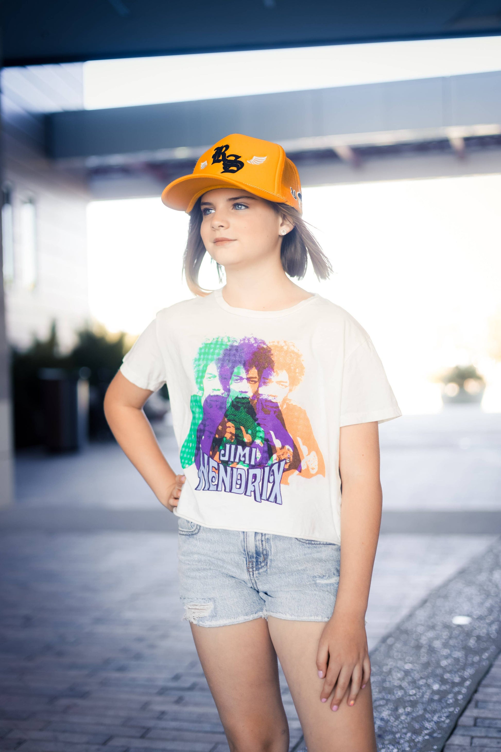 a child standing in a parking lot wearing denim shorts, a Jimi Hendrix rainbow crop top, and a yellow cap