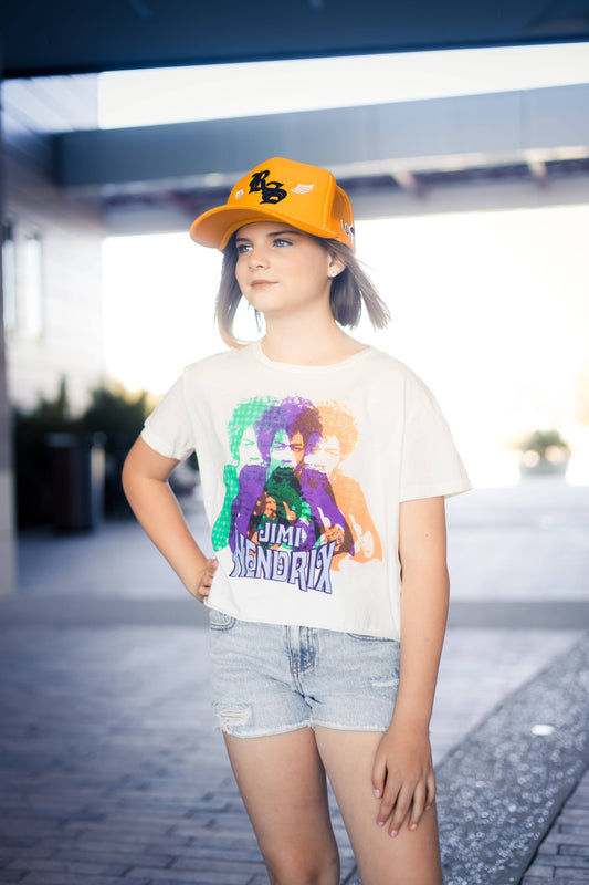 a child standing in a parking lot wearing denim shorts, a Jimi Hendrix rainbow crop top, and a yellow cap