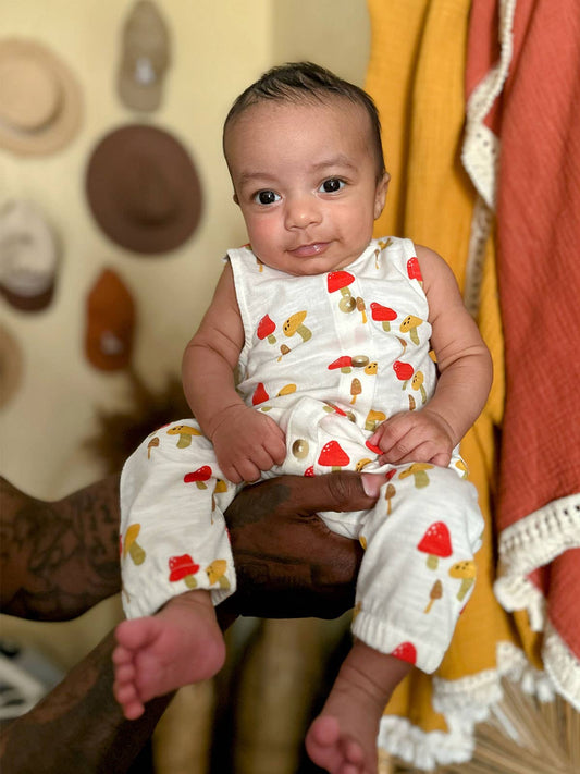 Baby wearing a white jumpsuit with a red and yellow mushroom print sitting on a grownup's hands