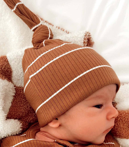 Baby wearing a brown knit hat with white stripes on a soft surface