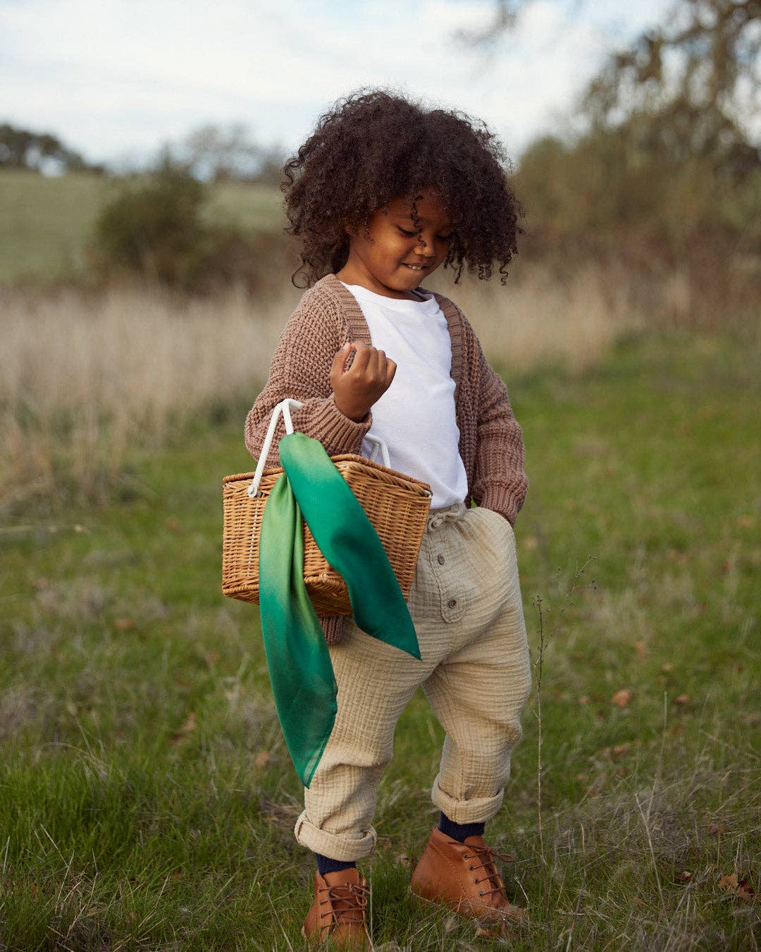 A child walking in a field holding a straw basket with a mini forest playsilk tied to the handle