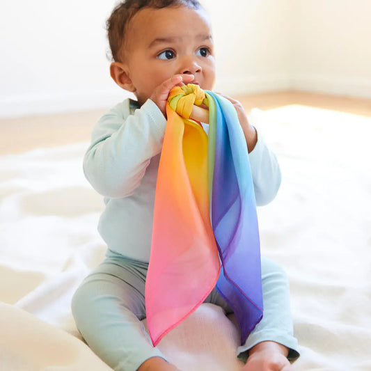 a baby sitting on a blanket chewing on a rainbow silk teether