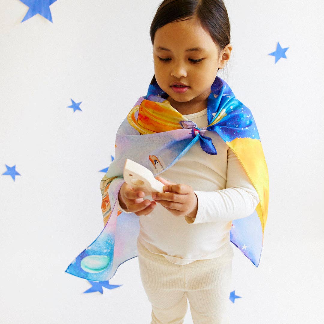 Child holding a wooden rocket wearing a space seek and find playsilk as a cape