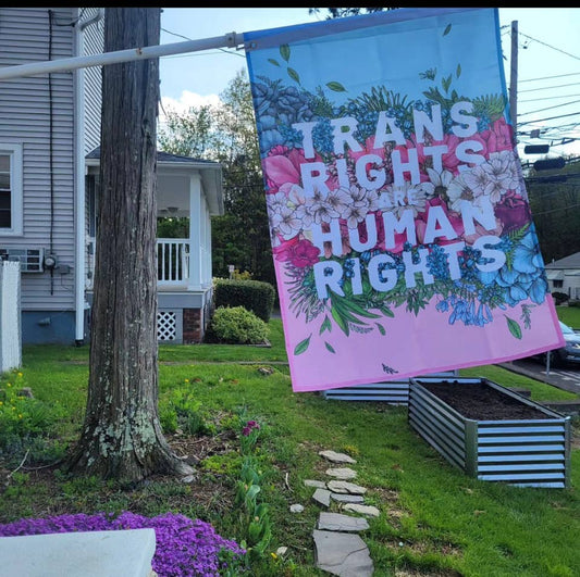 trans pride flag with a flower print decoration and the words "trans rights are human rights" printed on it hanging from a flagpole