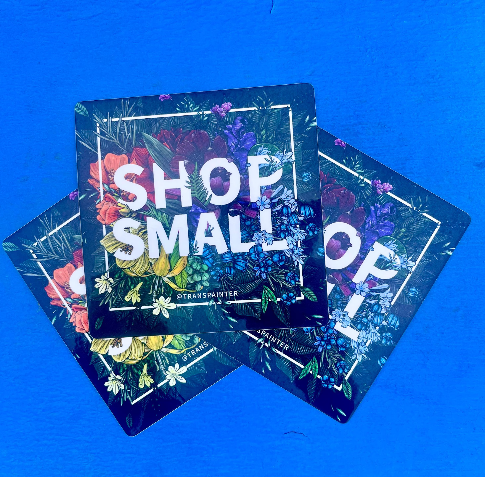 4x4 art by Transpainter stickers that read "Shop Small" on a black background surrounded by rainbow florals.