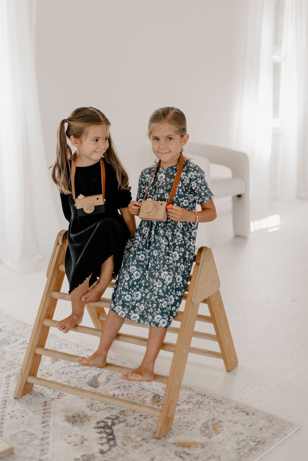 Two young girls sitting on a wooden ladder in a bright room.