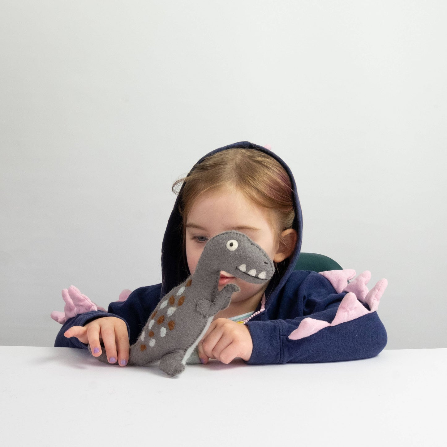 Child wearing a dinosaur-themed hoodie with a plush toy dinosaur in front of them on a white background
