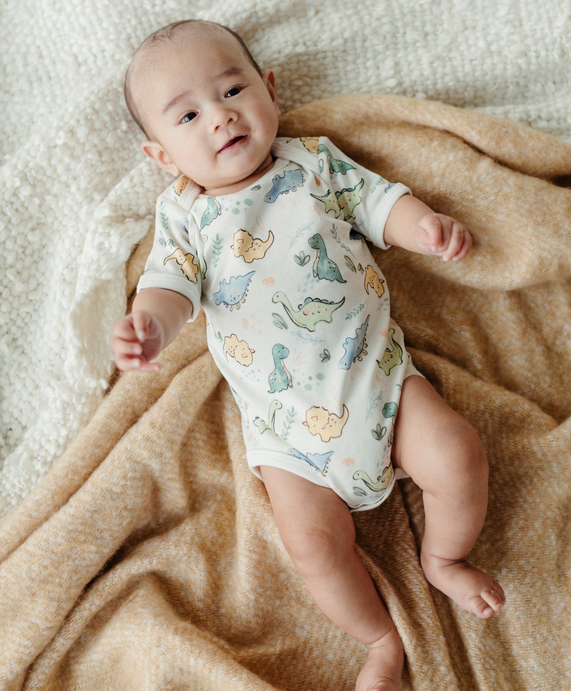 a baby laying on blankets wearing an organic short sleeve onesie with a dinosaur print
