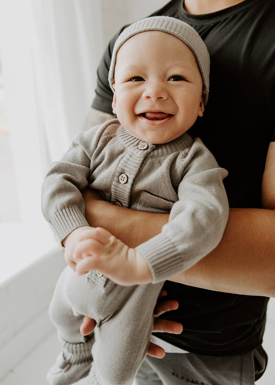 Baby in a gray romper and knit hat being held by an adult, smiling.