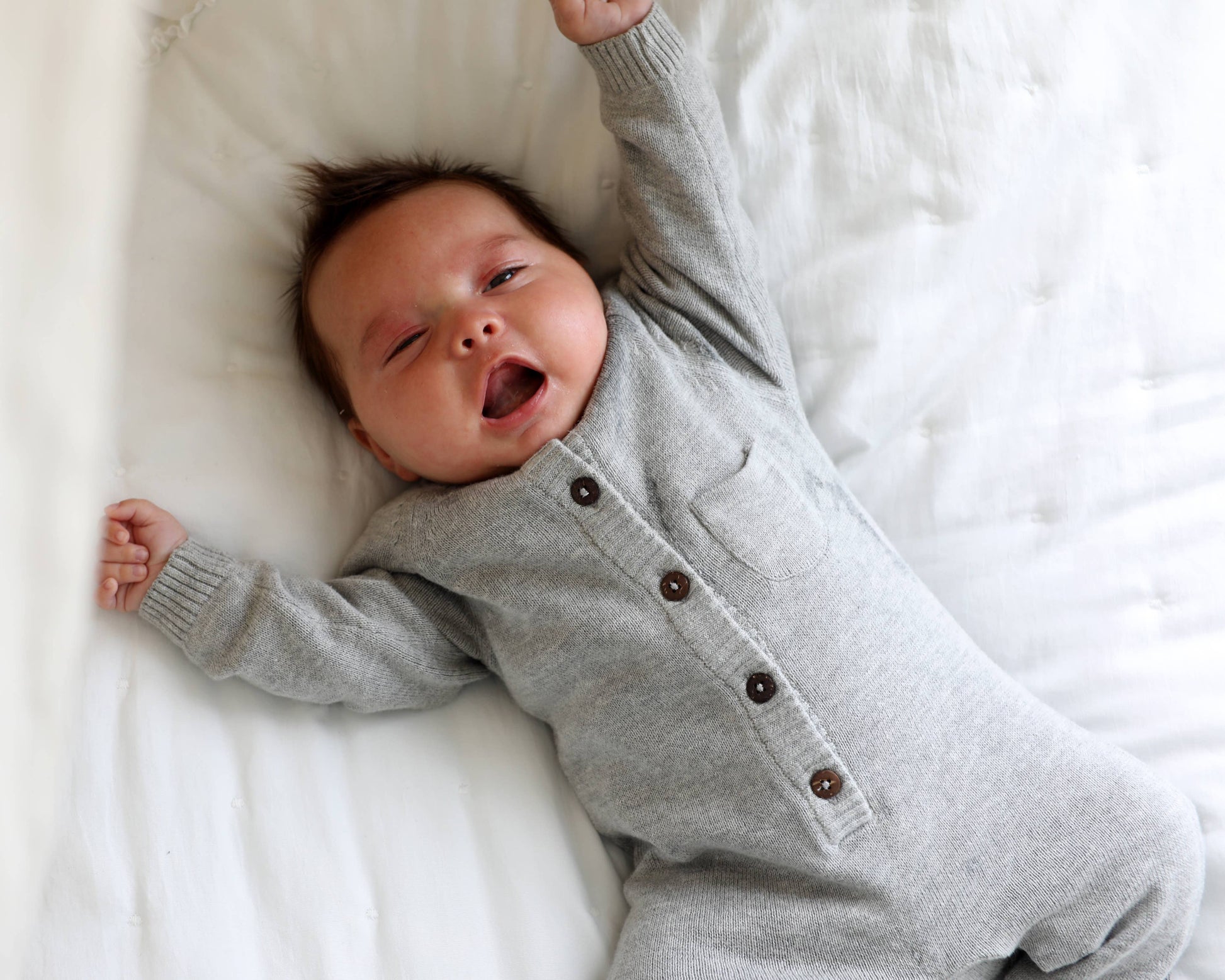 Baby wearing a gray romper with buttons, lying on a white bed.