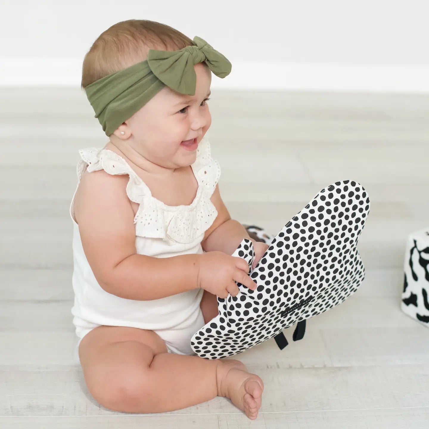 Baby sitting on the floor wearing a white outfit and green headband, holding a black and white polka dot crinkle toy