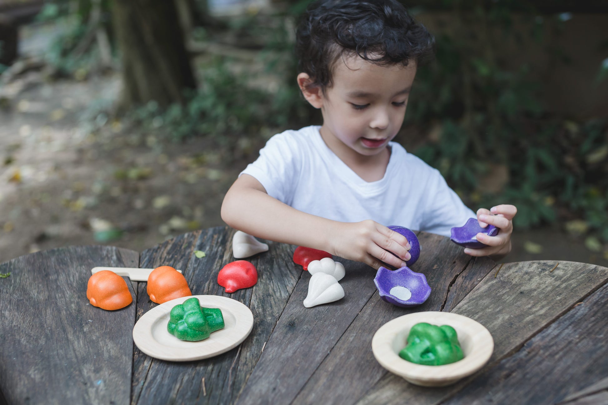A child playing with a wooden vegetable set on a wood table outdoors. the child has set the two halves of the broccoli on two wooden plates