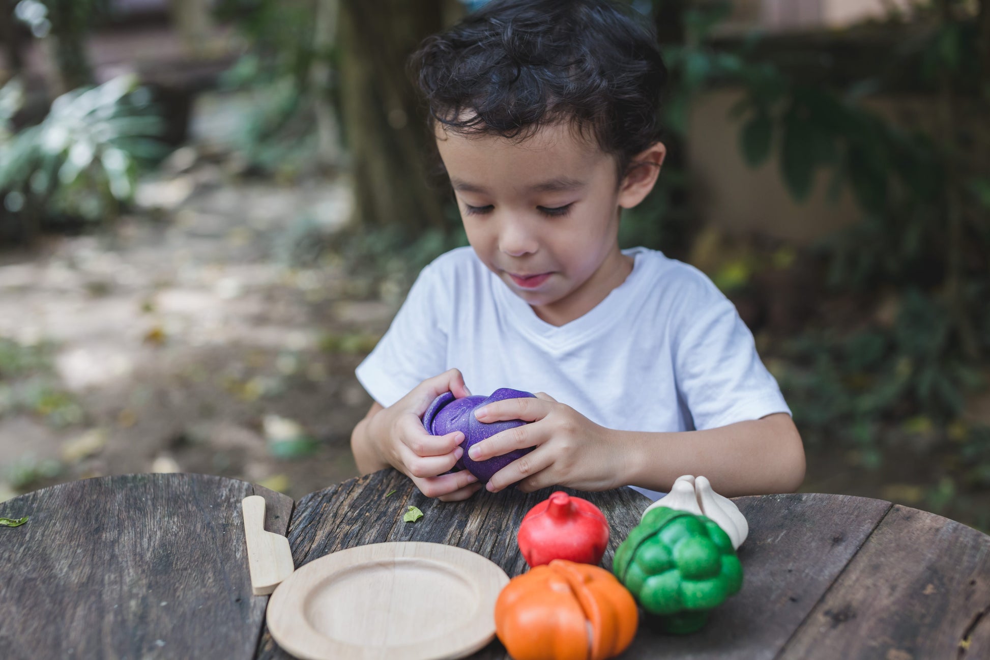 A child playing with a wooden vegetable set on a wood table outdoors. The child osopening the onion
