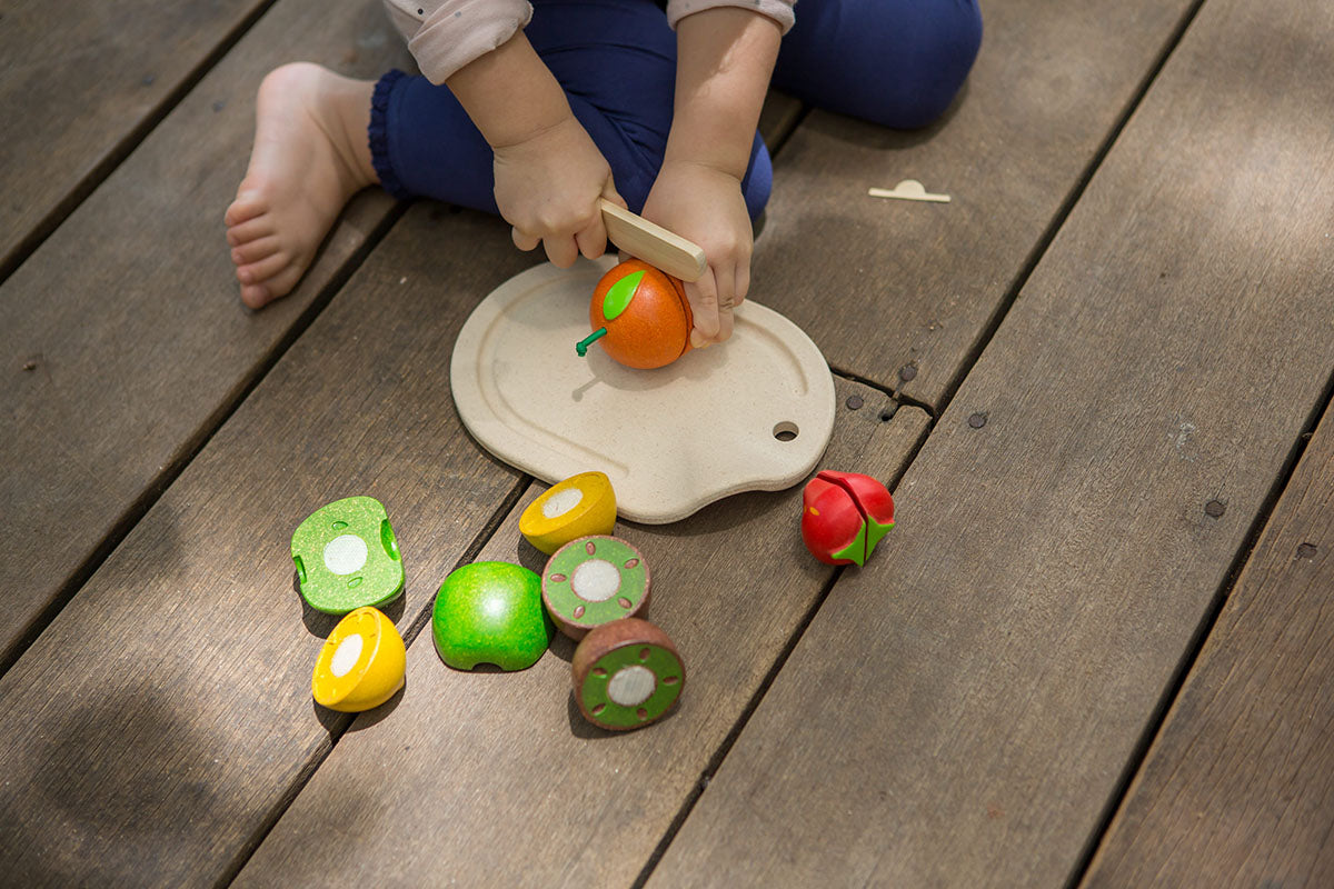 Child  outside on a deck cutting a wooden  velcro orange from the plan toys Assorted Fruit Set