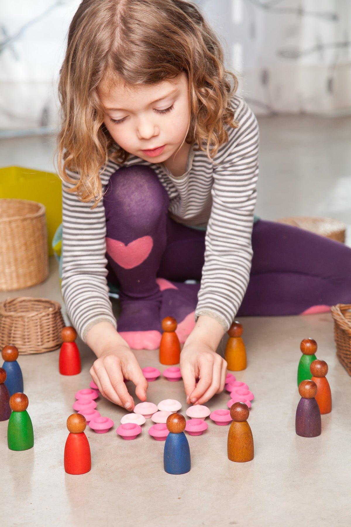 A child playing on the floor with colorful peg people, straw baskets, and a pink wooden flowers mandala.