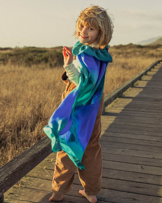 Kid on a wood board walk next to a grass field with a ocean silk cape on