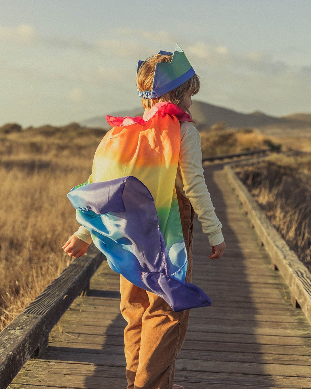 Child with rainbow cape and crown on a board walk with dried grass fields to the sides