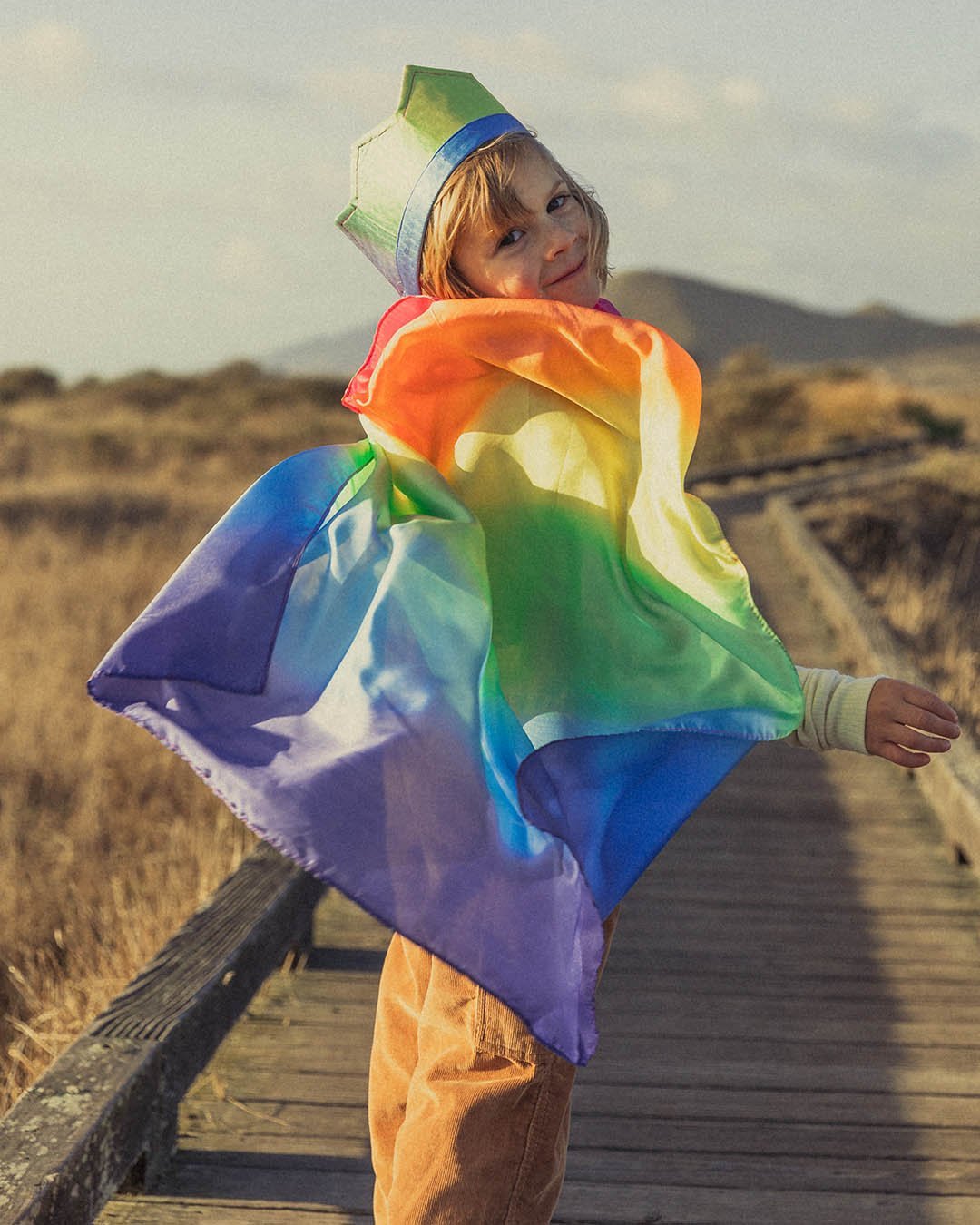 Child turning and smiling with rainbow cape and crown on a board walk with dried grass fields to the sides