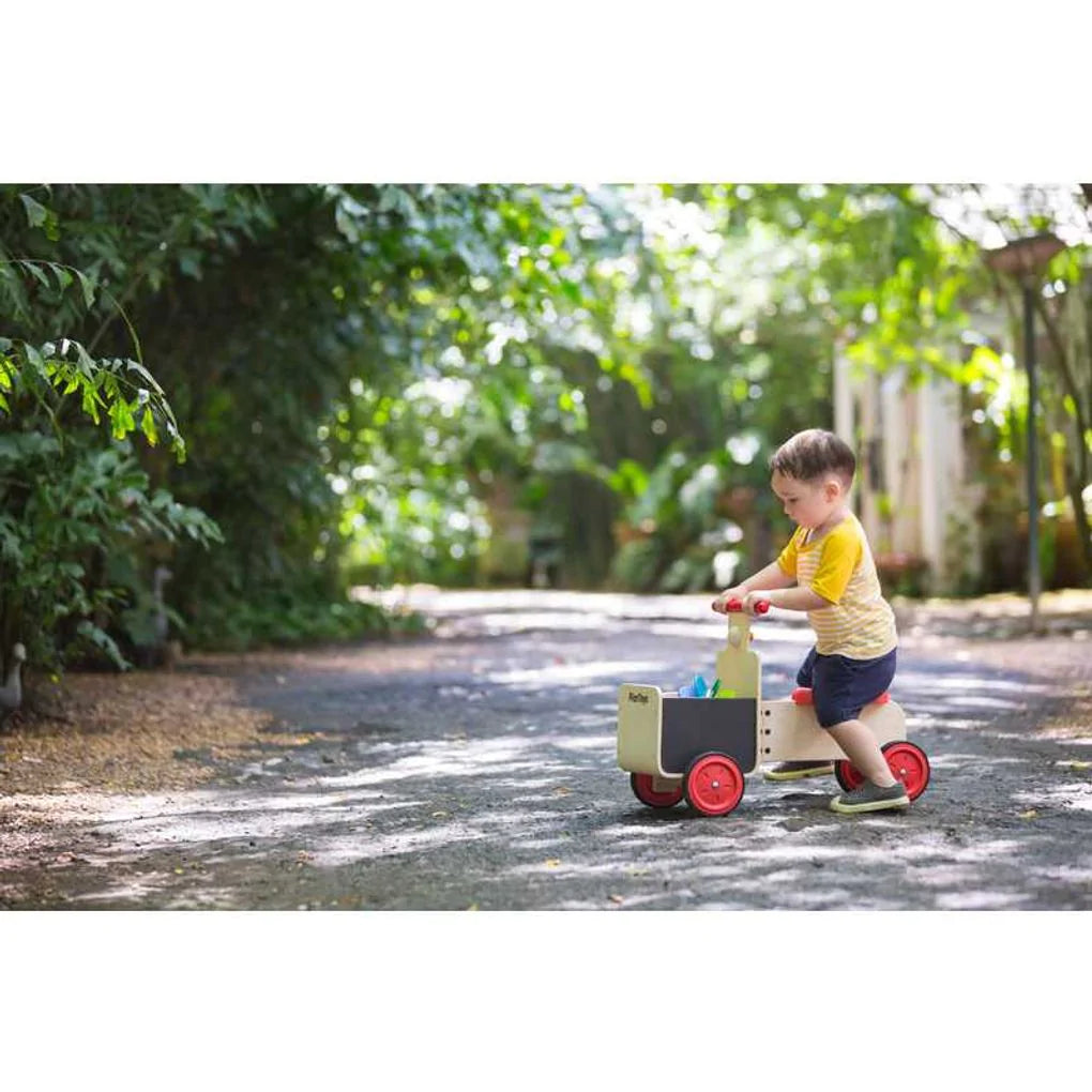 Child in a yellow and white shirt on a tree shaded road riding the plan toys wooden delivery bike