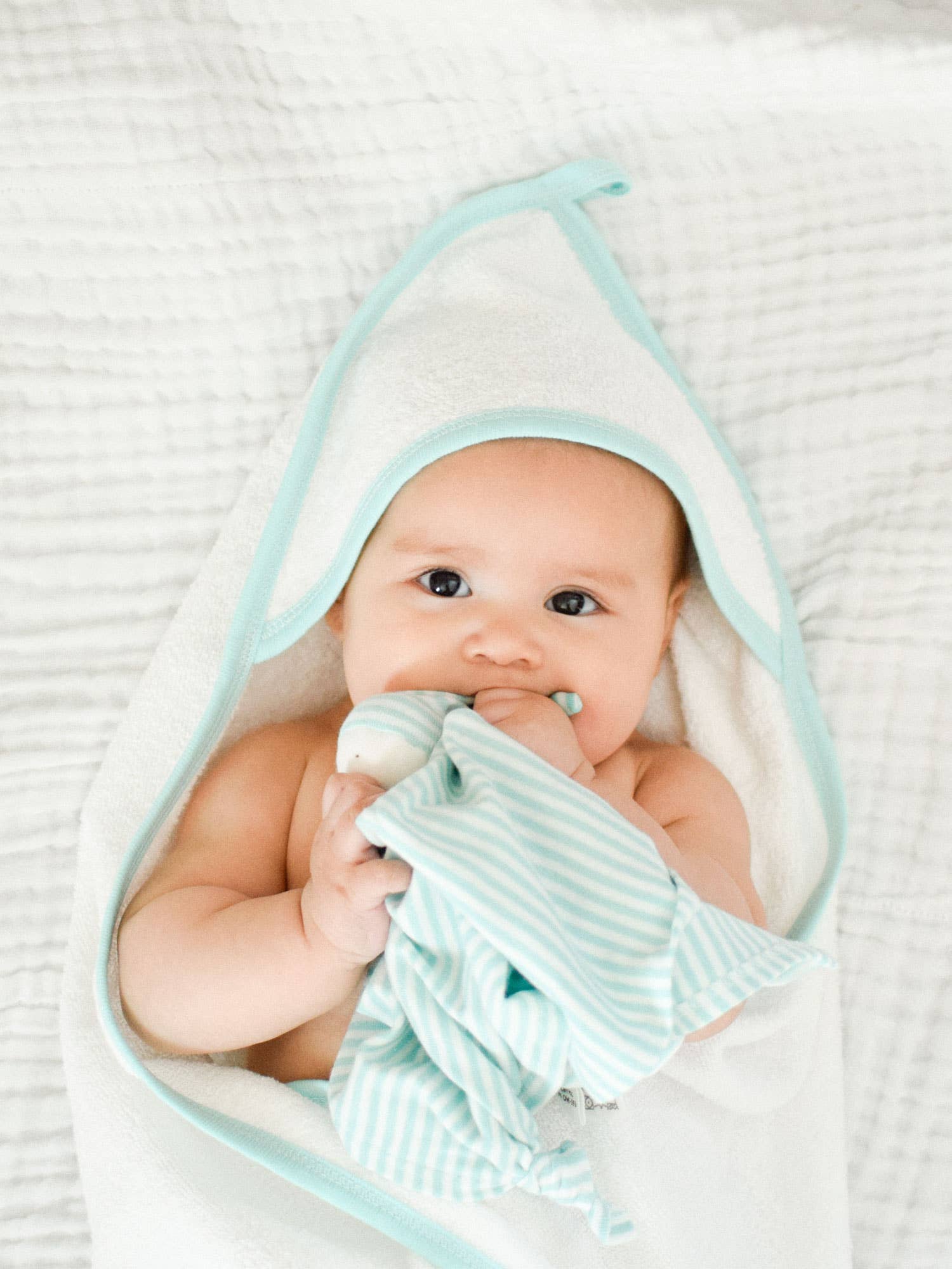 Baby laying on a white blanket wrapped in aOrganic Aqua Deluxe Hooded Towel