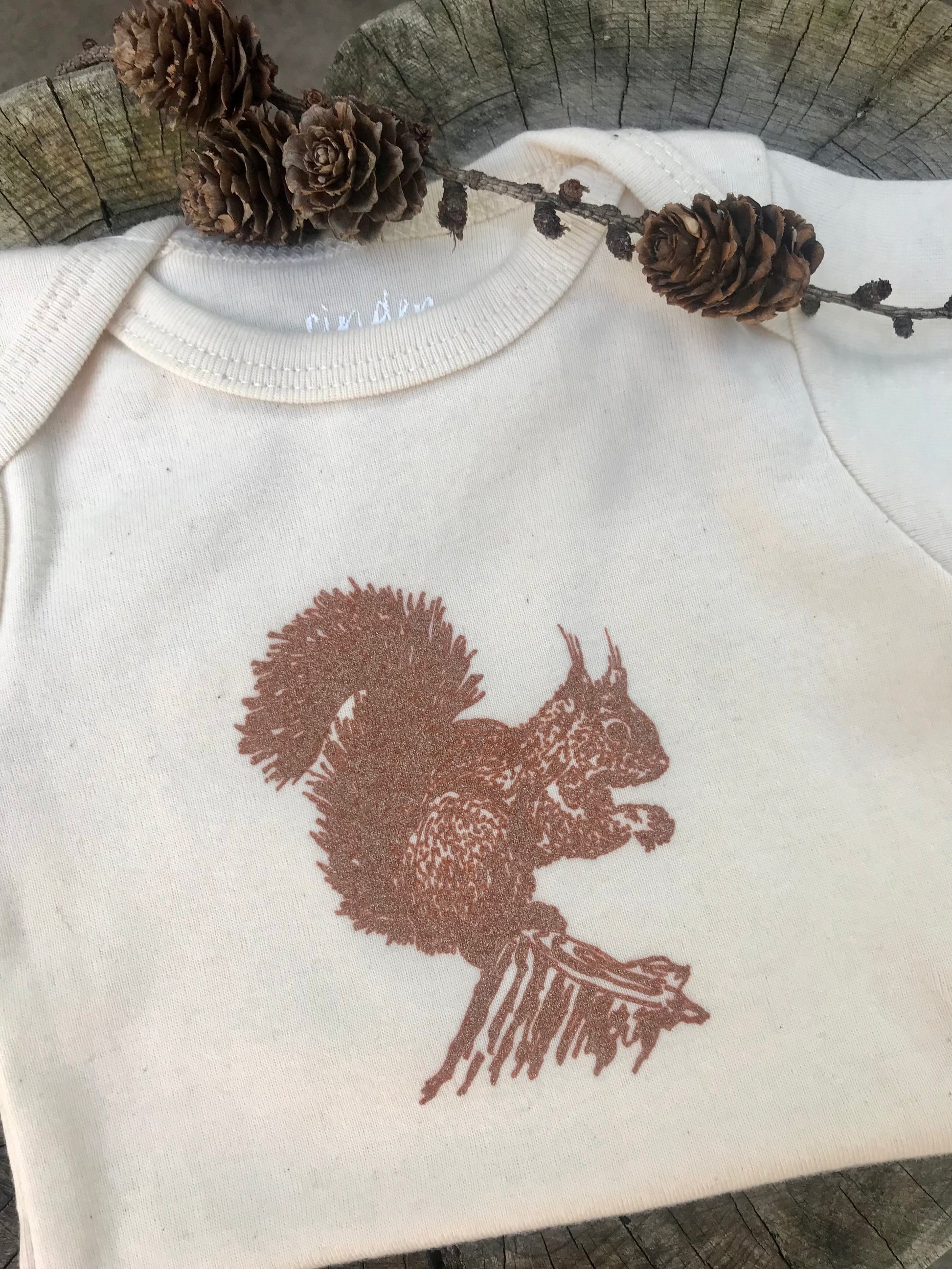 Squirrel Onesie folded on a tree log with pinecones