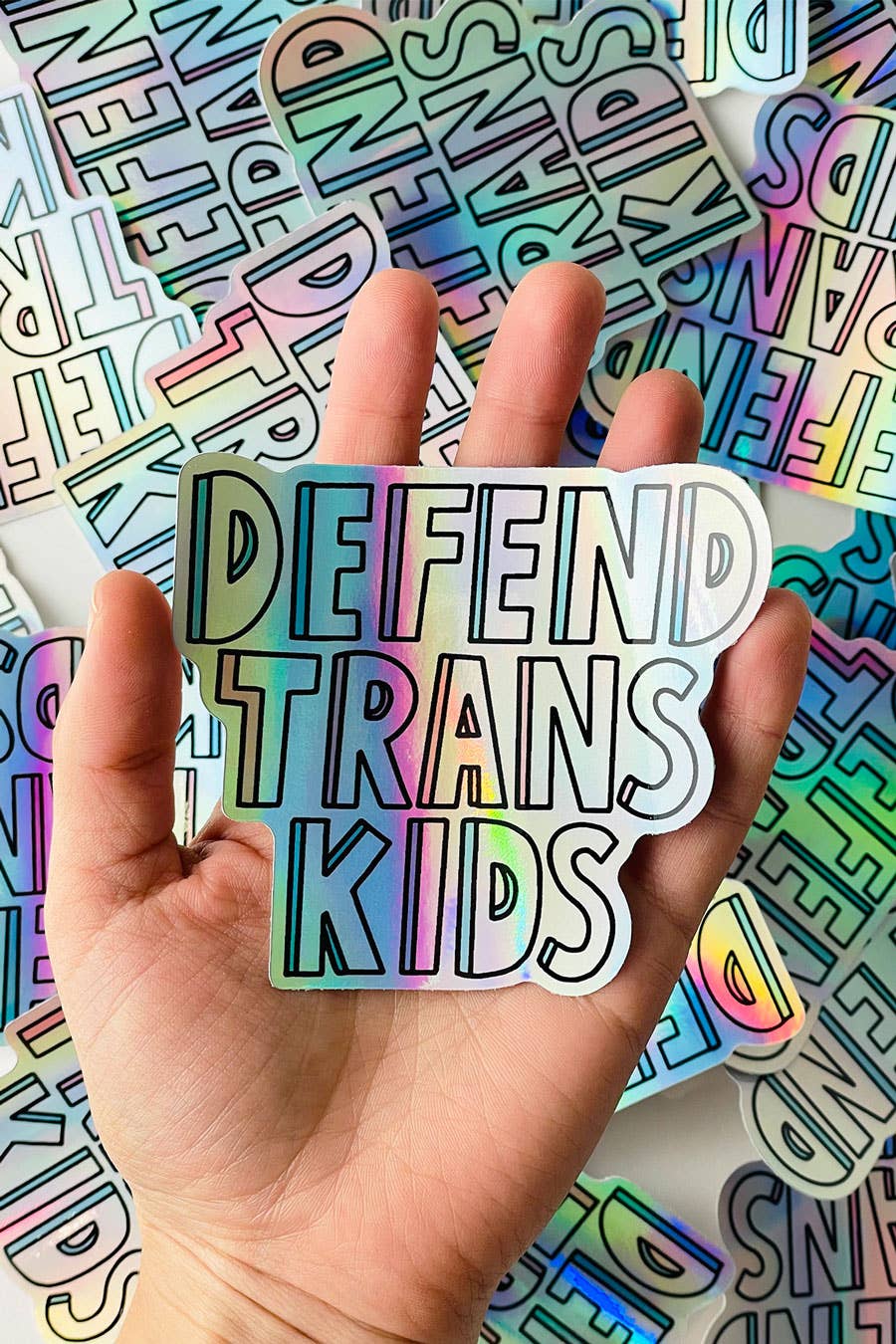 a hand holding a holographic vinyl sticker with the words "Defend Trans Kids"