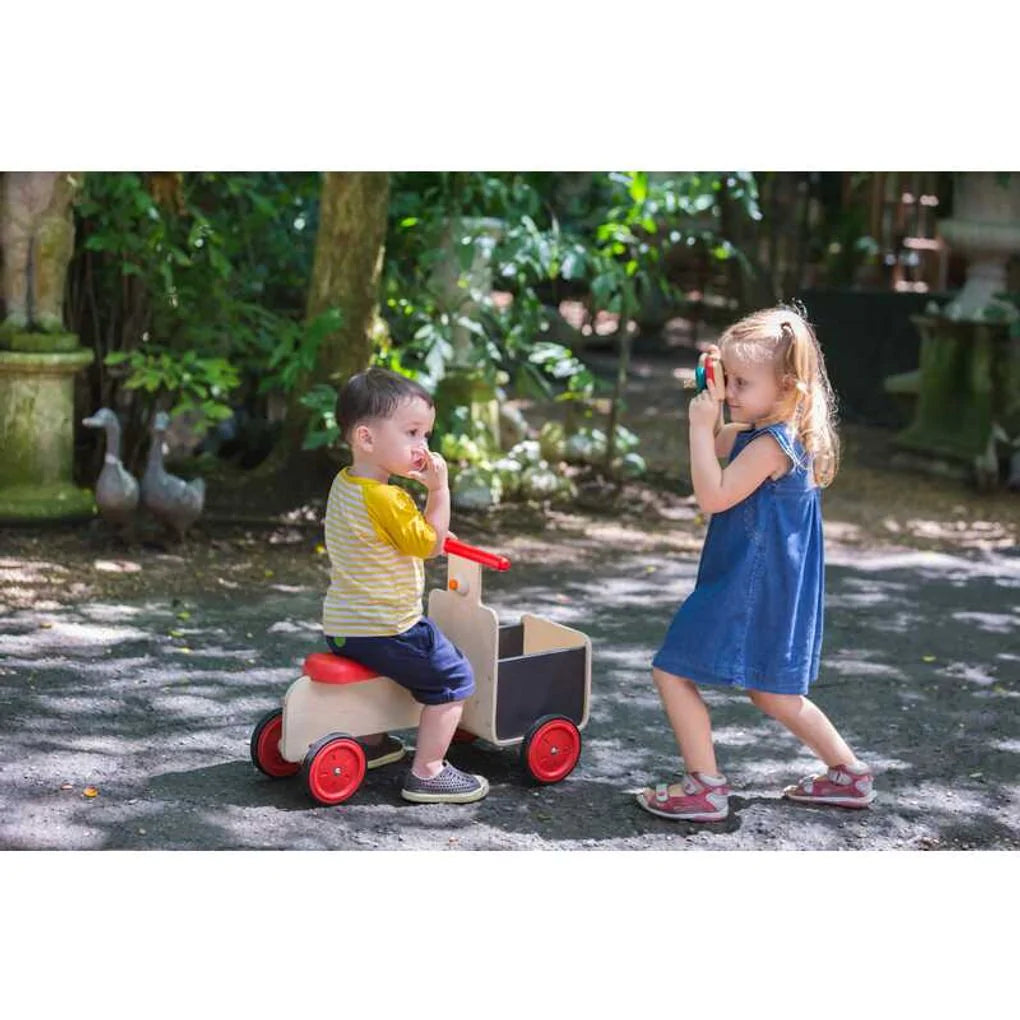 Children playing on a tree shaded road one child in a yellow and white shirt riding the plan toys wooden delivery bike and another kids in a denim dress using a toy camera