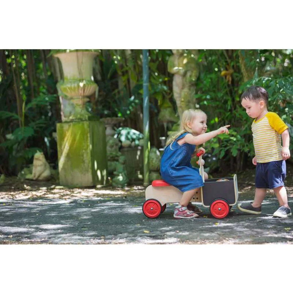 Kids playing in Sun dappled light child in a denim dress riding the plan delivery bike gesturing to another kids in shorts and a yellow and white stripe shirt